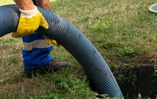 A worker from the local septic tank service holds a hose while standing near an open septic tank on a grassy area.