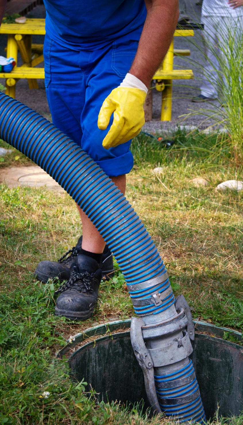 A person in blue work attire and yellow gloves operates a large hose, inserting it into a manhole on grassy ground, efficiently representing the expertise of Local Septic Tank Service.