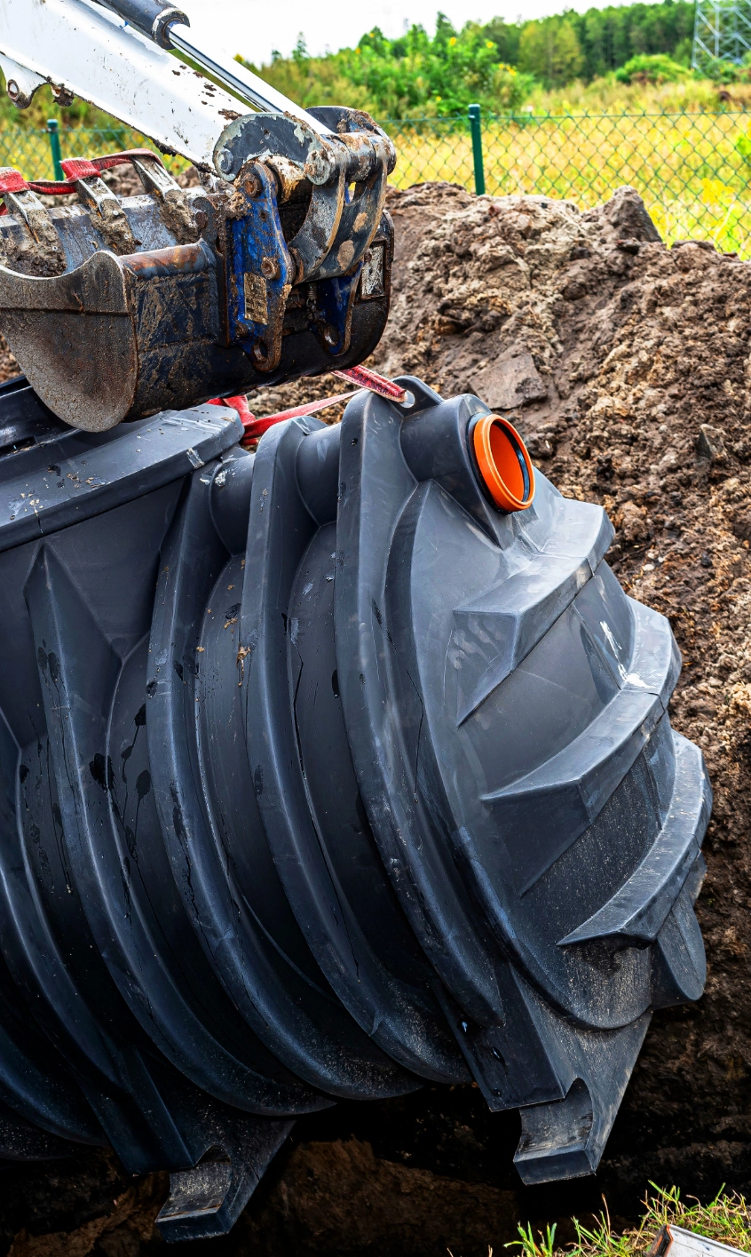 An excavator from a local septic tank service carefully places a large, grey, plastic septic tank into a trench at a construction site. Dirt piles and a grassy field are visible in the background.
