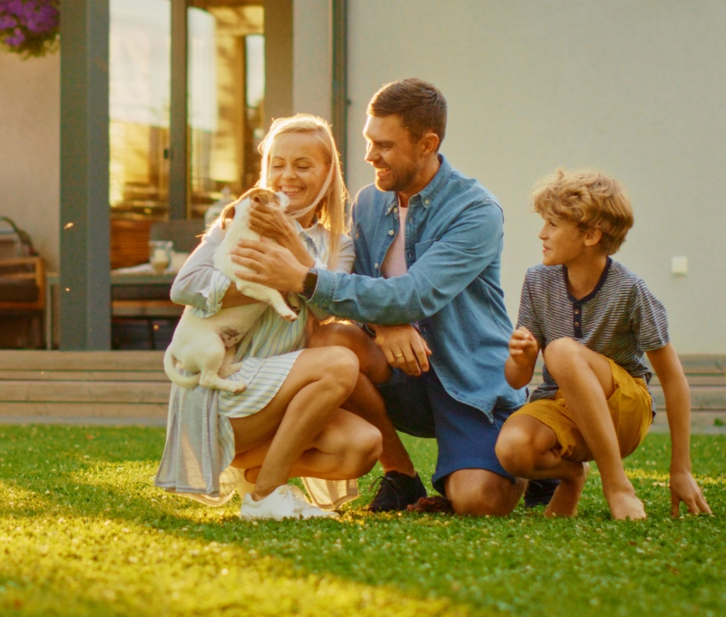 A woman, man, and child are kneeling on a grassy lawn, smiling and playing with a small dog in front of their house.