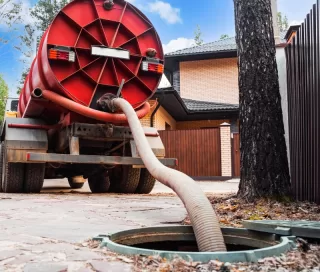 A large red tanker truck is parked near a tree with a hose extending from the truck into an open manhole, suggesting sewage maintenance or waste removal.