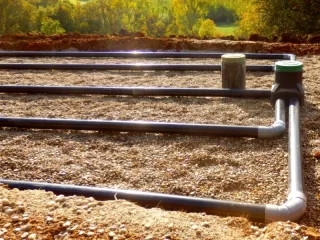 A drainage system featuring black pipes laid out in a gravel bed, outdoors, with green trees and sunlight in the background.