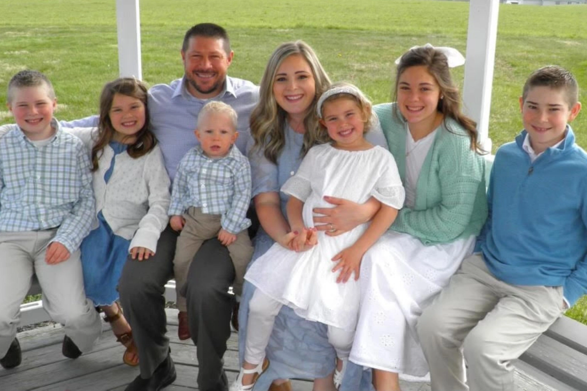 A family of eight, comprising three boys, three girls, and two adults, sits closely together on a gazebo bench in a grassy area while wearing coordinated outfits.